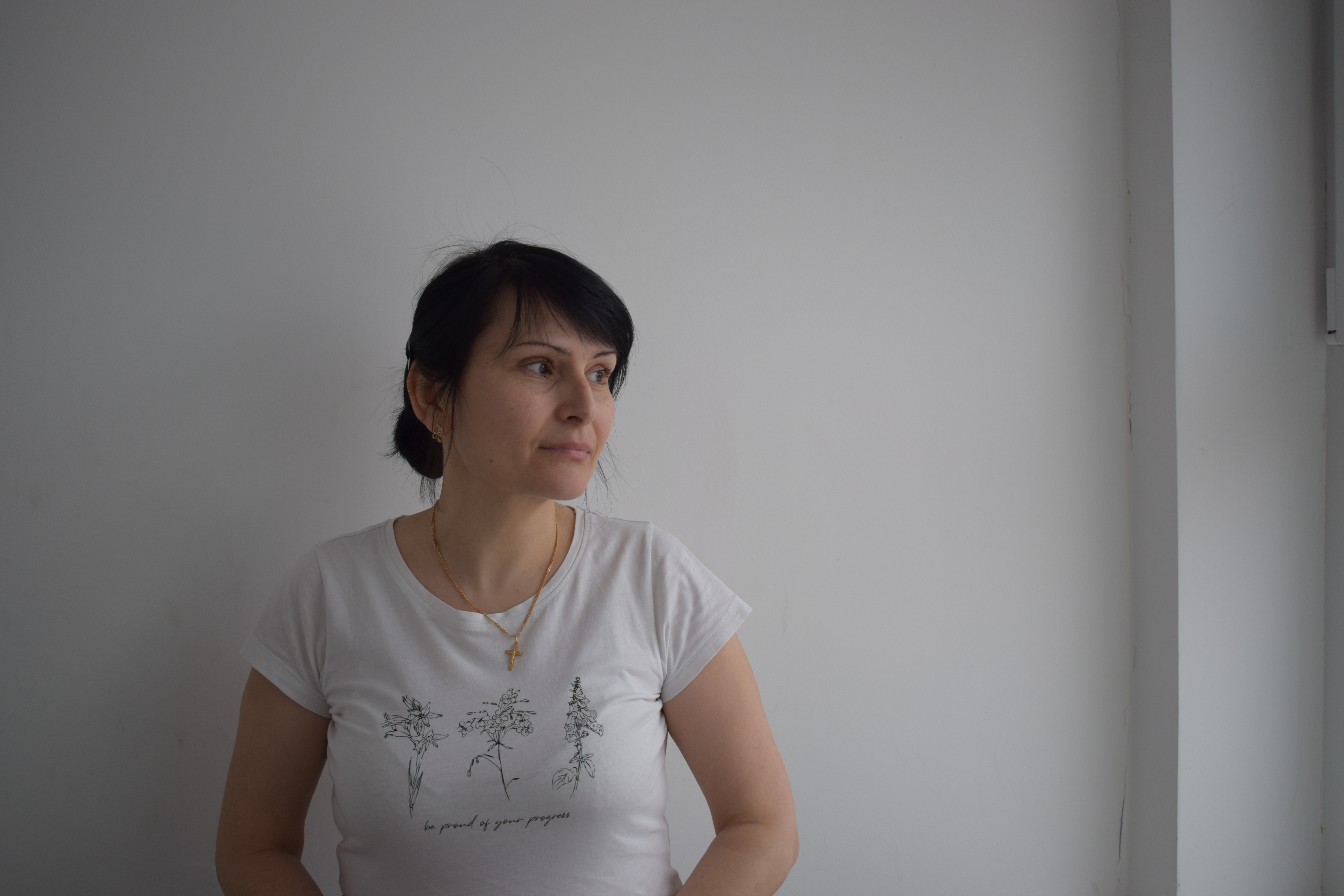 Kobieta w białej bluzce stoi na tle ściany, patrzy w bok, na twarzy ma delikatny uśmiech.