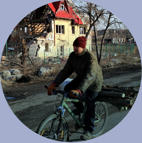 Mężczyzna zimą na rowerze wiezie drewno na opał w tle zrujnowany dom jednorodzinny, wschodnia Ukraina, 2015. Fot.: Jacek Marczewski