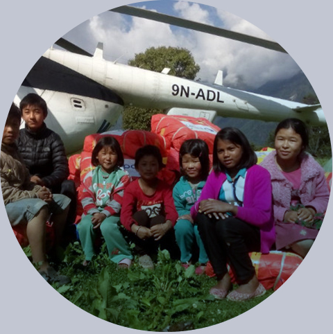 Grupa dzieci siedzi na paczkach z tarpolinami obok helikoptera, który dostarczył jako pomoc, PAH Nepal, 2015 rok. Fot.: PAH